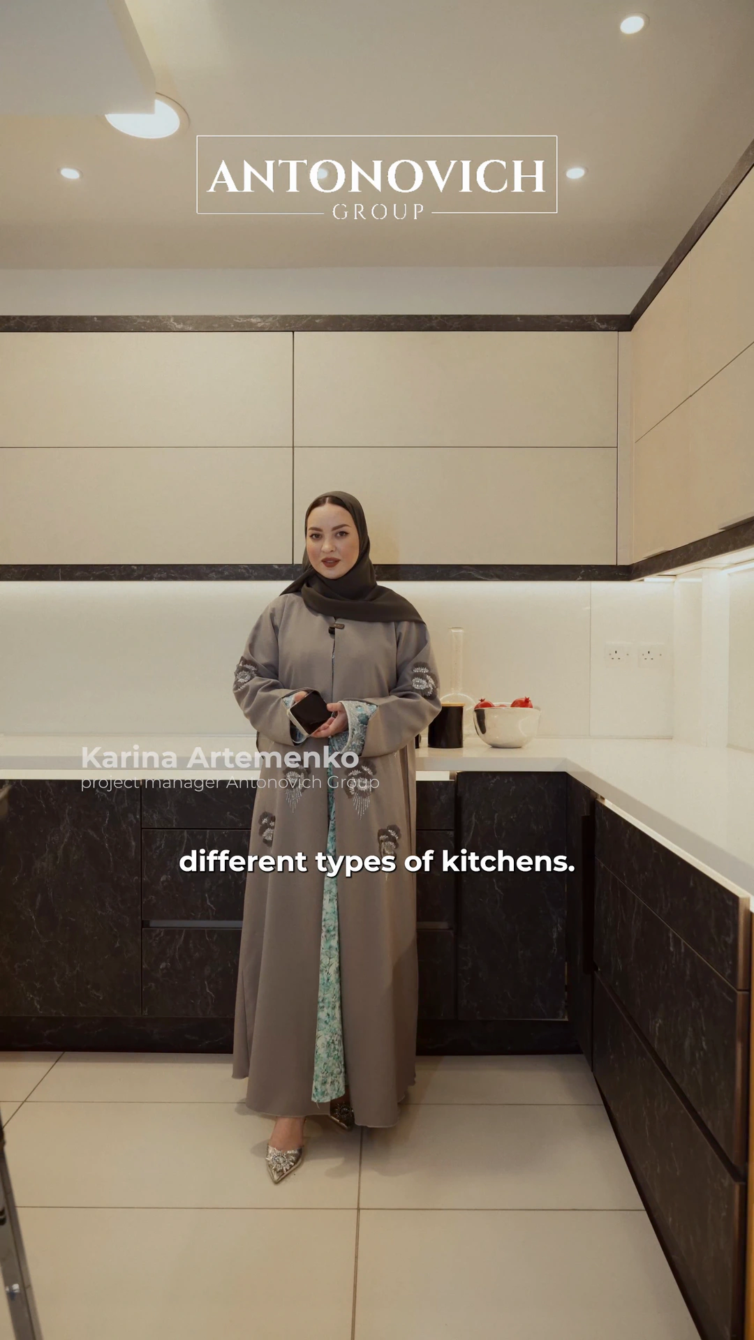 Presenter in a grey abaya showing a modern beige and black kitchen in the Antonovich Group showroom in Dubai