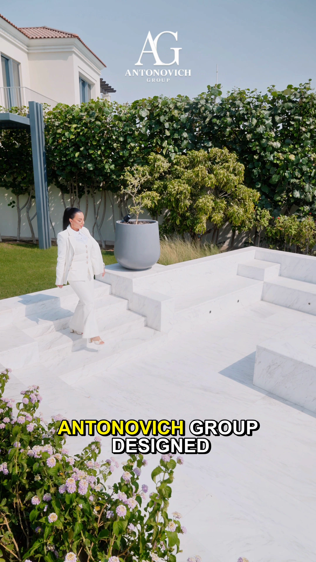 Woman in a white suit walking down white marble garden terraces with flowers and a green hedge, designed and built by Antonovich Group