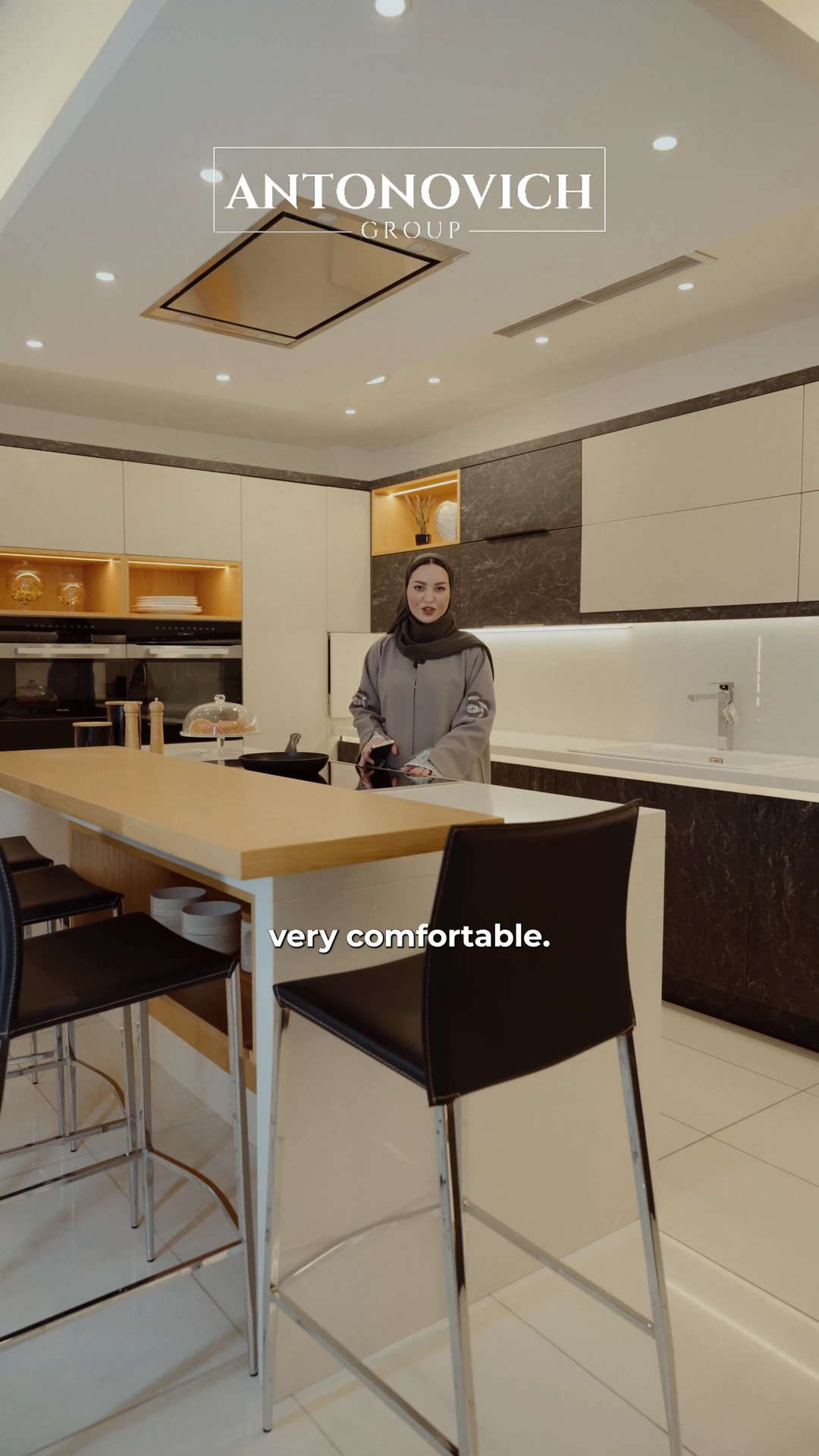 Presenter in a grey abaya showing cream and dark-wood kitchen fronts, pull-out storage and an island with bar stools