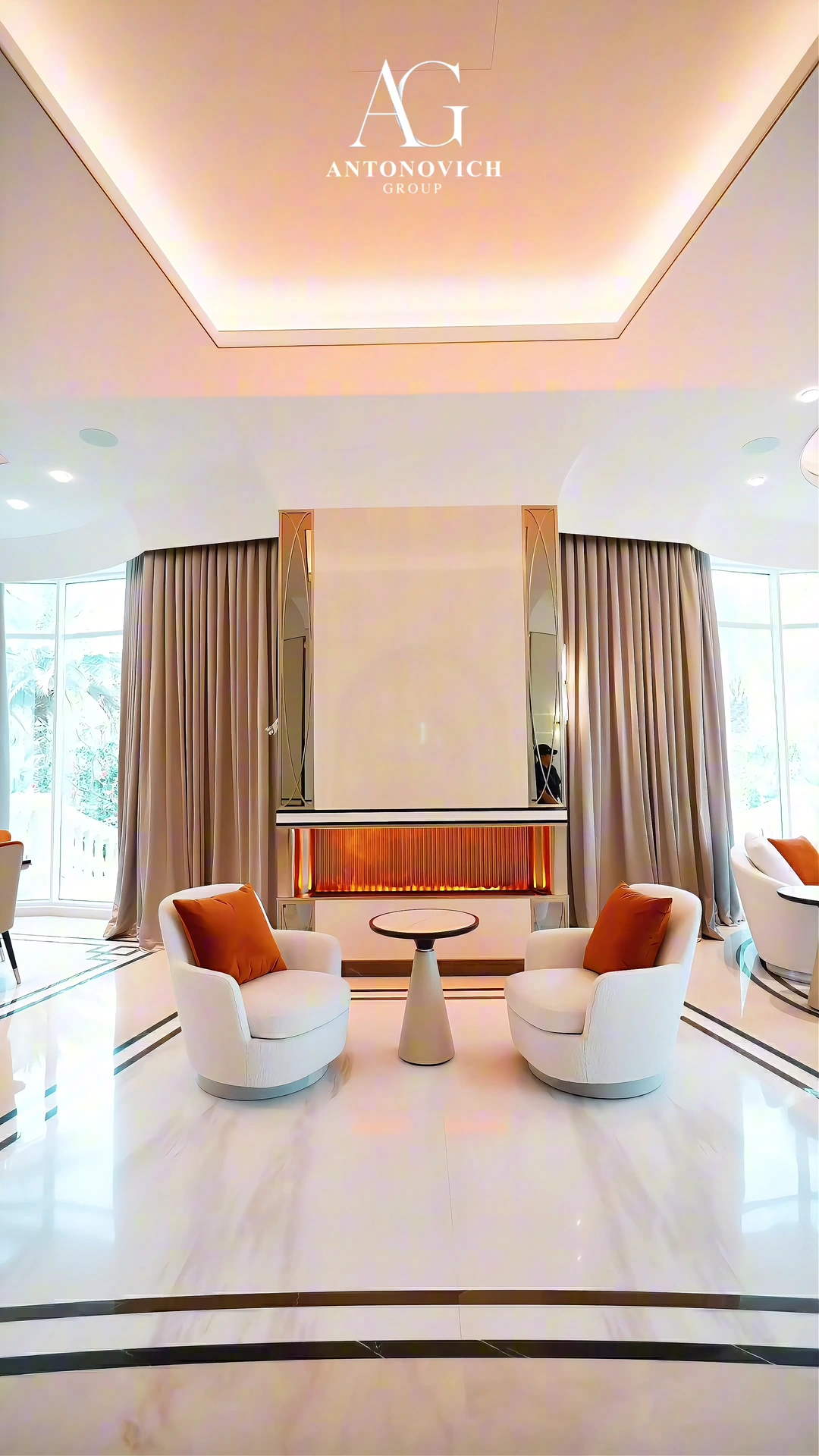 Renovated villa lounge in Dubai with two white armchairs and orange cushions either side of a round table, a linear fireplace in a fluted niche and a polished marble floor