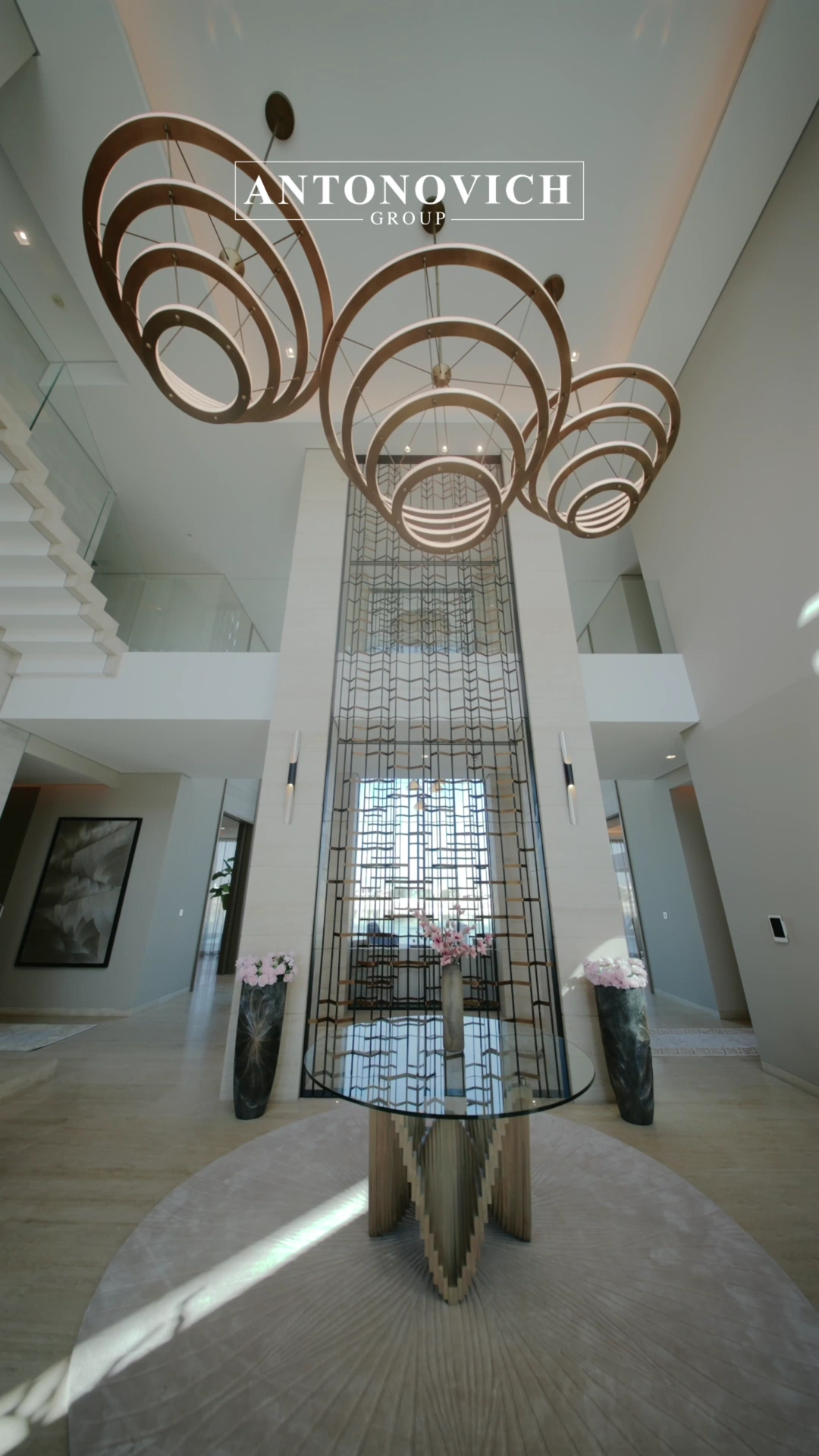 Double height entrance hall of a renovated villa on Palm Jumeirah with three bronze ring chandeliers, a round glass table holding pink blossom on a circular rug and a full height geometric metal screen