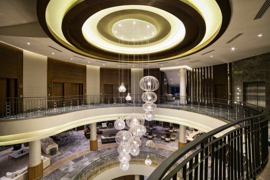 This picture shows two people having a picnic in a park. The woman is wearing a white shirt and pink skirt and the man is wearing a blue shirt and khaki shorts. They are sitting on a patterned blanket on the grass and have a basket of food between them. They are also surrounded by trees, plants, and bushes.