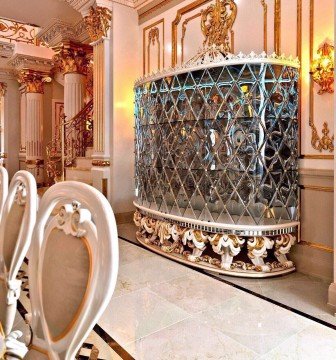 In the picture, there is a woman dressed in a purple and green sari. She is standing outside in front of a building. The woman has her hands folded in a prayer-like position and she is looking up towards the sky.