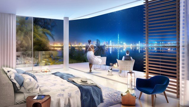 This is a picture of a white-tailed deer (Odocoileus virginianus) standing near some trees and grass in a forest. The deer is a medium-to-large sized animal, with a reddish-brown coat mottled by white spots. Its large ears are pricked up, indicating it has heard or seen something.