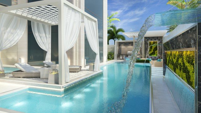 This picture shows a man standing near a waterfall. He has his hands in his pockets, and appears to be looking off into the distance. The waterfall is cascading down from a rocky cliff surrounded by lush green vegetation. The sky is blue, and the sun is shining brightly.