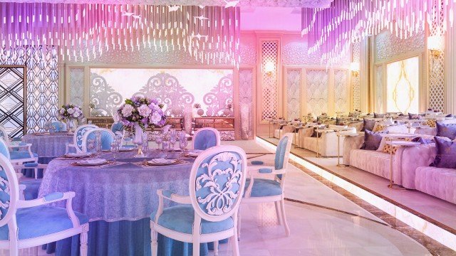 The picture shows a man in a business suit and mask, standing in front of a white background. He appears to be holding a laptop in his hands, with the screen open, showing a pile of gold coins.