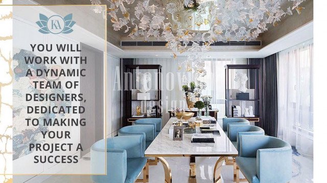This picture shows an ornately decorated marble staircase with gold fixtures and a black banister, situated in a grand foyer. The staircase leads up to a landing with large curtained windows where two people are standing. The walls of the foyer are covered with intricate patterned wallpaper and house numerous pieces of art.