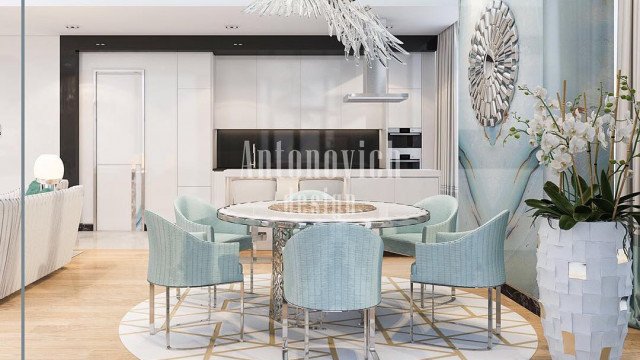 This picture is of an ornately decorated modern living room. The walls are painted in a light grey, with large geometric gold-painted designs along the edges and around the windows. There is a white fireplace in the corner of the room, and in the center of the room is a round black coffee table surrounded by four velvet upholstered armchairs. The furniture is accented with small gold accents, and the room is finished off with two floor lamps and a chandelier for lighting.