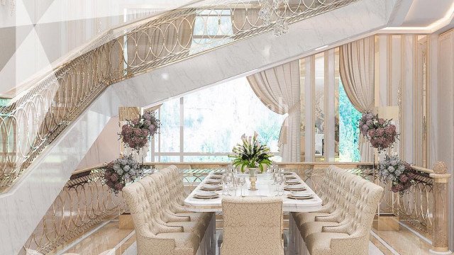 This picture shows a luxurious modern living room designed by Antonovich Design. The room is decorated in shades of white and gold, with a large ornate mirror over the fireplace and a floor-to-ceiling window that looks out onto a balcony or patio. The furniture is plush and inviting, and there is a rich dark wood flooring. The chandelier and lighting fixtures provide a warm and inviting atmosphere.