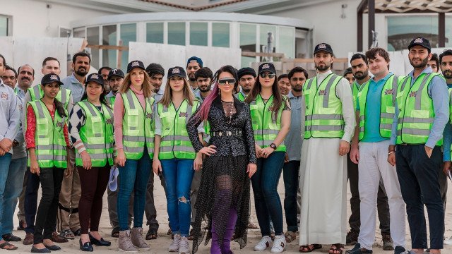 This picture shows an interior design featuring pastel colors and a modern design style. The room has a light blue wall with white trim, striped gray and blue curtains, a bright pink sofa with white throw pillows, and a round coffee table with a white top. The floors are light hardwood, and the walls are adorned with artwork and shelves. A large potted plant also adds a bit of greenery to the space.