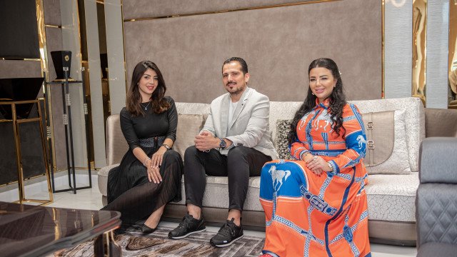 This picture is of a luxurious bedroom designed by Antonovich Design. The room is decorated in soft colors, with beige walls and white furniture. The furniture includes a four-poster bed, two night stands, a dresser and mirror set, an armchair, and a chaise lounge. There is also a large window with sheer curtains and a patterned rug in the center of the room.