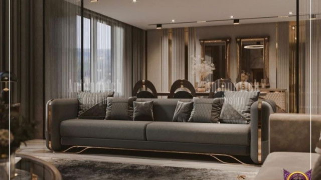 This picture shows a luxurious master bedroom interior. It has a large bed with a beige headboard and white bedding, a long bench at the foot of the bed, two contemporary armchairs in a darker beige, and several accent pieces like a tall lamp, artful sculptures, and vases. The walls are painted an elegant light grey and the carpet is beige.