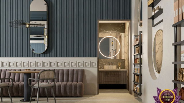 This picture shows an interior design concept from Antonovich Design. The space features a large area rug, white walls, and a mix of traditional and contemporary furnishings. There is a fireplace with dark wood and cream-colored marble accents, two windows with light sheers, and several armchairs in various shades of beige. In the background there is a built-in bookcase with various books and a tall curtained window. There are also a few pieces of art hung on the wall. The entire room has a bright and inviting atmosphere.