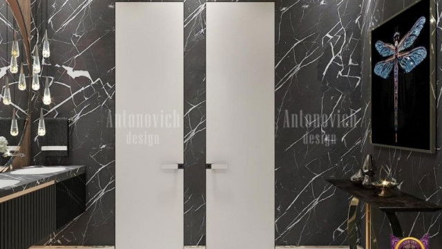 This image shows a luxurious and modern bedroom with grey velvet furniture and a mirrored accent wall. The bed has a light grey velvet headboard, adorned with a large bedspread and multiple round pillows in shades of grey, beige, and white. In front of the bed, there is a wooden nightstand and a light grey velvet armchair with a tufted back. The focal point of the room is the mirrored wall behind the bed, which reflects the entire room, giving it a greater sense of depth and spaciousness.