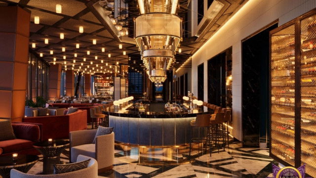 This picture shows a luxurious interior of a kitchen space. The room features a large island with white marble countertops that extend outward on both sides, and features a stainless steel range in the center. A white glass chandelier hangs above the island, adding light and elegance to the room. On the right side of the kitchen, there is a large built-in refrigerator and pantry area with a glass front door and a wooden countertop. The kitchen also includes wooden cabinets and light-toned marble flooring throughout.