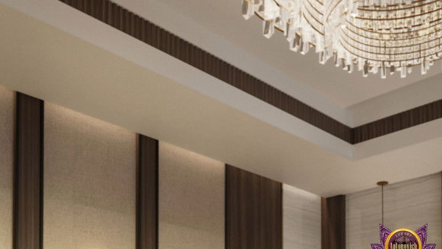 This picture shows a modern interior living room design with sleek and minimalistic furniture, incorporating an off-white sofa, a glass coffee table, and two gray velvet armchairs. It also features a large television mounted to the wall in a black wooden frame, a round work desk, and two decorative plant pots placed on either side of the room. The white walls provide a crisp, bright backdrop for the wood-colored floor and the pieces of furniture.