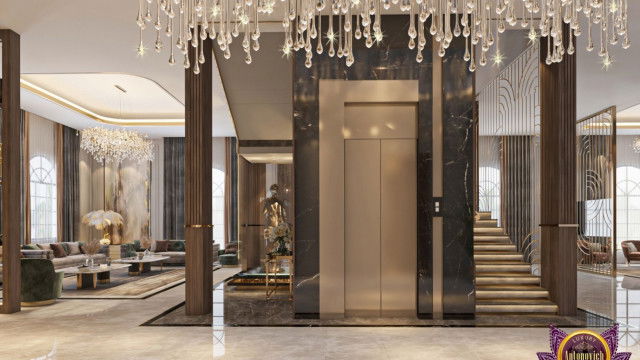 This picture depicts a modern, luxurious living room designed by the interior design firm Antonovich Design. The room features a tufted white patterned sofa with several decorative pillows, a white fur heirloom rug, and a large sculptural centerpiece that stands atop a marble-like table. On the walls are framed pieces of artwork and there is an intricate crown molding along the ceiling. A floor to ceiling window offers plenty of natural light and allows for views of the outside.