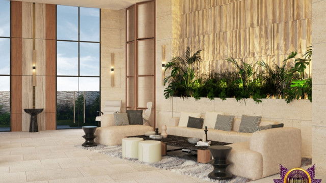 This picture is showing a contemporary living room with vaulted ceilings, featuring a curved sofa and two armchairs in a neutral color scheme. The walls of the room are painted white and adorned with modern artwork. A round dark wooden coffee table sits in the center of the space, and a blue and white patterned rug grounds the area. The floor-to-ceiling windows allow for plenty of natural light and provide a view of the outdoors.