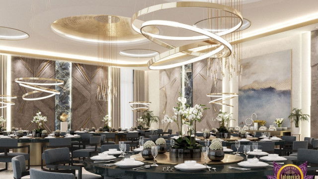 This picture shows a luxurious living room designed by Antonovich Design. It is decorated with modern furniture, gold accents, and crystal chandeliers. The walls are painted in a light grey color, and the floor is covered with a cream-colored rug. There is an ornate fireplace on one wall, and the opposite wall features a large, abstract art piece that adds a touch of sophistication.