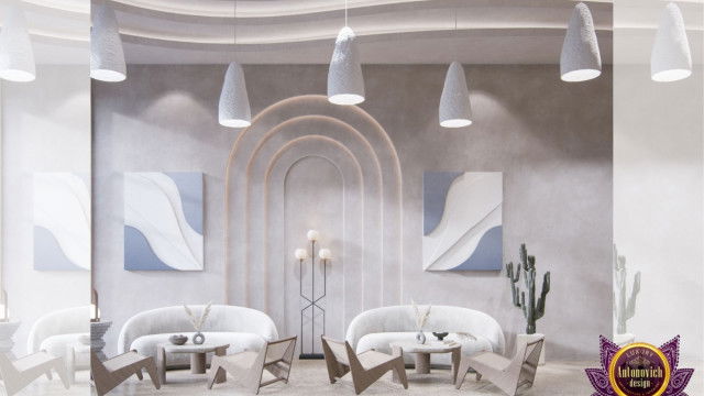 This picture shows a luxurious interior design featuring an open-concept living and dining space. The main focal point appears to be the large white custom-built media wall in the center of the room that houses a state-of-the-art home entertainment system. The furniture is modern and plush, with a velvet maroon sofa and accompanying leather armchairs. The overall look is one of opulence and sophistication, beautifully finished off with hints of gold accenting the ceiling and wall fixtures.