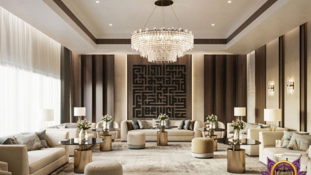 This picture shows a modern luxury living room with a pale grey and beige color palette. The focal point of the room is the large, dark grey velvet sofa, which is situated in front of a tall, contemporary fireplace with a black marble surround and hearth. An ebony grand piano stands against the far wall near a large window covered by sheer curtains. On either side of the sofa are two tall armchairs in coordinating fabrics, and the entire room is accented with gold and glass decor pieces.