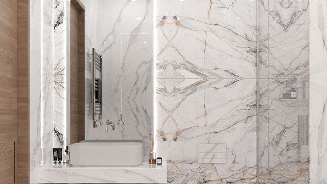 This picture shows a modern interior design of a living space. The space is lit by several recessed LED lights throughout the ceiling and walls, and features a white L-shaped sofa with colorful throw pillows featuring geometric patterns. There is a tiled fireplace to the left, set against a charcoal gray accent wall with a large rectangular artwork suspended above it. To the right is a curved wooden bar counter with barstools and a variety of glasses, liquor bottles, and other barware. The flooring is a light hardwood.