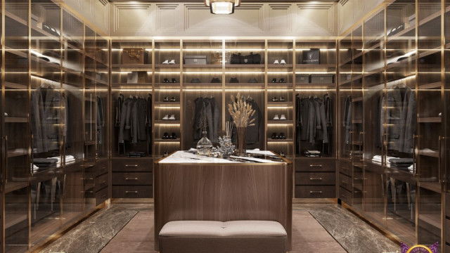 This picture shows a modern bedroom with sophisticated design elements. There is a white bed with tall grey and black headboard, and a white and grey linens. On the sides, there are two nightstands with table lamps and accessories. The walls are painted in a light grey color and on one wall there is a marble accent with a large abstract painting. Above the bed, there is an ornate mirror framed with a black and silver leaf pattern. The floor is laid with a grey carpet and there is a dark rattan armchair in the corner of the room.