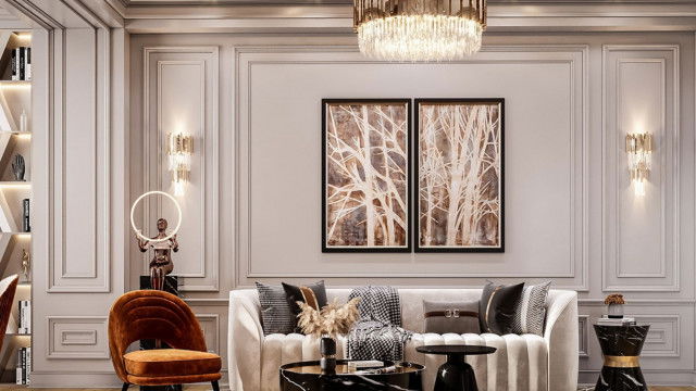 This picture depicts a modern apartment living room, with a sleek and minimalistic design. The room has white walls and furniture, contrasted by elements of dark wood, grey upholstery, and a few touches of greenery. There is an armchair in the corner and a sofa facing a large flat screen television. In the center of the room is a low coffee table where several stylish decor items have been artfully arranged.