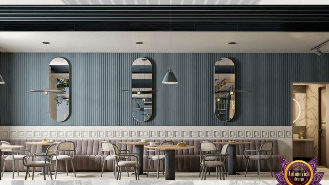 This picture is of a contemporary-style living room. The walls are painted light grey, and a dark grey accent wall stands behind the white leather sectional sofa. The room features a glass coffee table and an armchair in black upholstered fabric. There is also a round mirror and an abstract painting on one wall. The floors are a light wood, and there are several plants throughout the room, adding a touch of greenery.