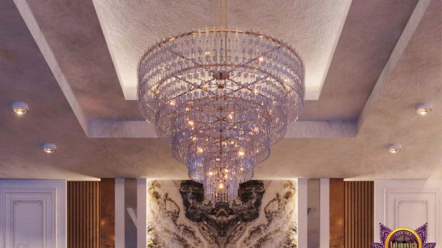 This picture depicts a modern luxury living room featuring neutral colors and clean lines. The room features an upholstered chaise lounge, a white coffee table on a furry rug, an ottoman, and an oversized armchair with gold accents. There is also a large abstract painting on the wall and an ornamented ceiling light that adds to the elegance and sophistication of this luxurious space.