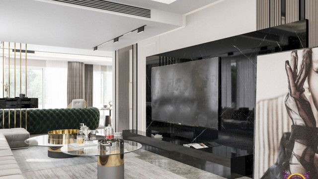 This image shows a luxurious living room with an elegant and classic design. The room features a white marble fireplace with a tall mirror above, a brown leather couch and armchair across from the fireplace, a glass coffee table between the two, an ornate rug in the center of the room, and two tall bookshelves with ornately-framed artwork on either side. There is also a tall white window treatment with sheer curtains, a crystal chandelier, and large vases with plants on either side of the fireplace.