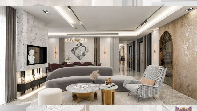 This picture shows a luxurious sitting room in a modern home. The room features a large, curved white sofa along with a round white ottoman in the center of the room. The walls are painted a light shade of blue and are adorned with multiple pieces of art. A striking crystal chandelier hangs from the ceiling and provides light to the entire room. There is also a large fireplace on the wall across from the sofa and several potted plants around the room.