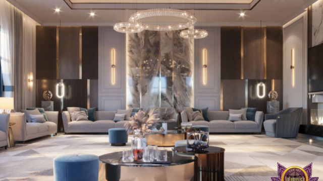 This picture shows a luxurious modern living room designed by Antonovich Design. The walls are covered in a light gray velvet fabric, with a beige and gray floral patterned area rug laid out in the center of the room. A large gray and white sectional sofa is arranged against one wall, with two dark brown armchairs and a low glass circular table placed nearby. The room is illuminated by several modern glass pendant lights, and the back wall is adorned with a large abstract painting.