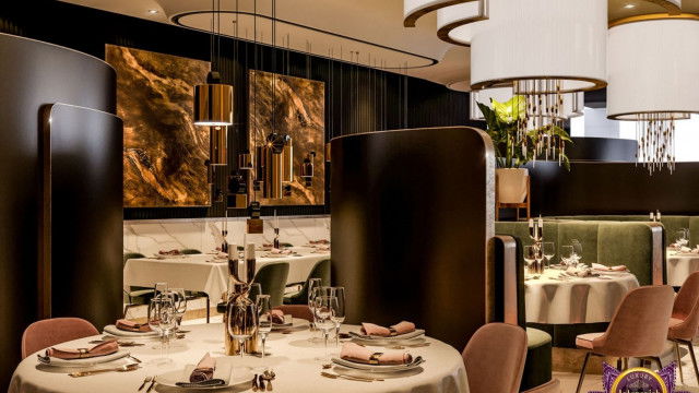 This picture shows two elegant and modern chairs in a luxurious living room. The chairs are cream in color, with tufted detailing along the back. They have slim gold legs and armrests and are placed near a large glass table. The backdrop consists of grey curtains, dark stained wood walls, and a large mirror framed in gold metal.