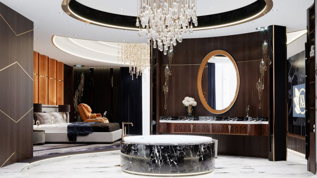 This picture shows a luxurious bedroom interior design. The walls are painted in soft gray with modern wallpaper accents that feature large white geometric shapes. A stunning chandelier hangs from the ceiling, while two sleek armchairs with matching ottomans provide comfortable seating near the window. The room features a grand wooden bed with a plush tufted headboard and matching bedding. Stylish artwork decorates the walls, and a contemporary bench is situated at the foot of the bed.
