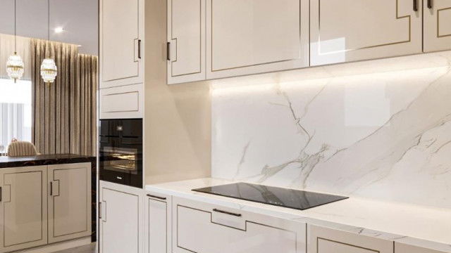 This picture shows a luxurious living room with a large sectional upholstered in white velvet. The walls are light grey in color and feature several paintings and photographs. There are two small mirrored accent tables near the couch, and a large glass coffee table in the center. To the left is a large window with golden curtains, overlooking a stunning view of the city skyline. The room is accented with luxurious touches such as a crystal chandelier and golden lamps.