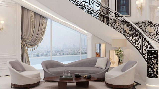 This picture is of a modern and luxurious living room designed by Antonovich Design. It features high ceilings, floor-to-ceiling windows on one side, a fireplace, and comfortable plush seating. The walls and furniture are decorated in shades of gray, while the flooring is light hardwood. There are also elegant wall hangings and artwork that add to the room's aesthetic.