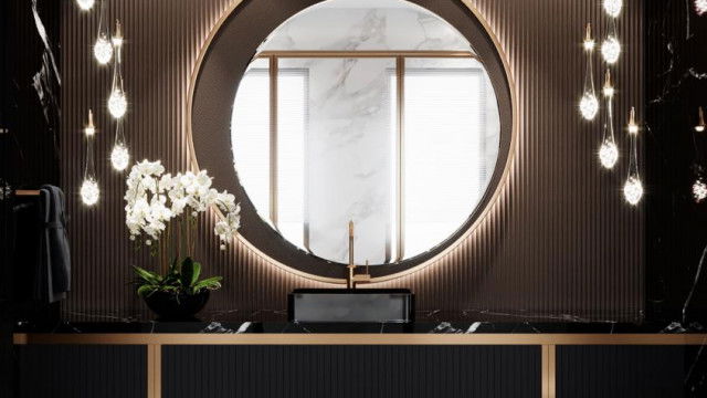 This picture shows a luxurious gold-accented bathroom. There is a free-standing bathtub in the center with a dark brown wooden base and a beige tiled exterior. On either side of the tub are two large, arched windows with white shutters that let in plenty of natural light. The walls of the room are decorated with an ornate white wallpaper and a large gold-framed mirror hangs in the center. There is a modern sink beneath the mirror with two tall gold taps and a marble countertop. Finally, the flooring is a light grey marble with