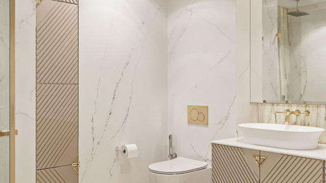 This picture shows a luxurious bathroom with beige and gold marble flooring, white walls, intricate gold-trimmed mirrors, and a gold chandelier. There is a porcelain corner tub in the center of the room, surrounded by wooden cabinets and several plants that bring life and color to the space. A white shower curtain hangs from the wall next to the corner tub, and a plush beige rug lies on the floor.
