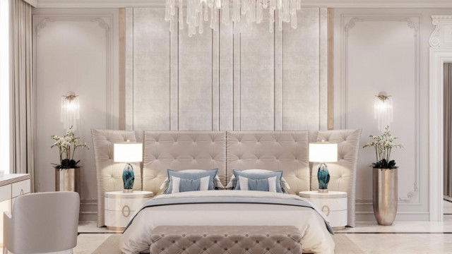 This picture is of a luxurious living room designed by Antonovich Design. The room features high ceilings with multiple elegant glass chandeliers, an ornately decorated fireplace, and a large area rug that gives the space a warm, inviting atmosphere. The walls are decorated with large canvas paintings and detailed moldings, and the furniture includes two sofas, an armchair, and two round tables. Numerous side tables and lamps provide additional lighting and decor.