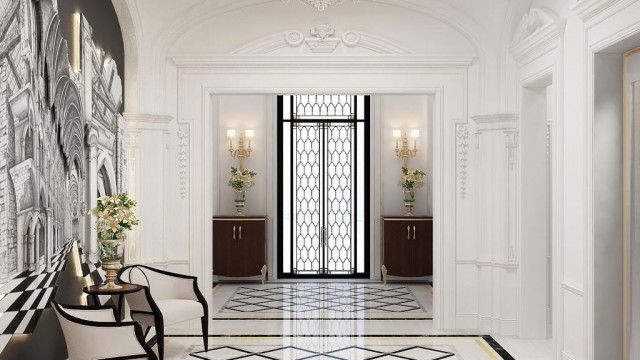 The picture shows a luxurious, modern bedroom with an elegant white and beige color scheme. The focal point of the room is the large four-poster bed which stands on an ornately patterned rug in shades of beige and brown. The walls are decorated with an eye-catching mural depicting an idyllic beach scene. Other features of the room include a built-in set of cabinets and drawers, a chaise lounge chair, and an upholstered armchair. A crystal chandelier hangs from the ceiling, providing a touch of glamour to the space