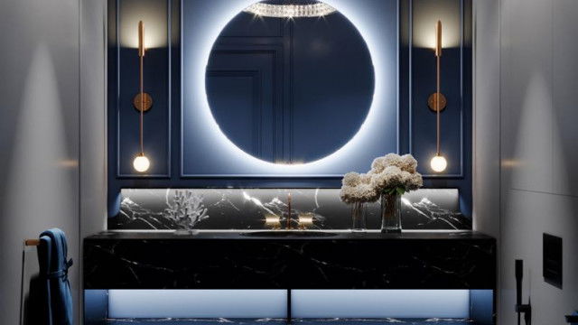 This picture is of a modern art deco style living room. The walls are painted in a deep, neutral color with a lighter shade of the same tone used for the accent wall. A light gray sofa sits in the center of the room with patterned throw pillows and a coordinating armchair to the side. A glass-top coffee table stands in front of the sofa, along with a round white side table and a floor lamp. A beige-colored rug is centered in the room, and two abstract art pieces hang on the walls.