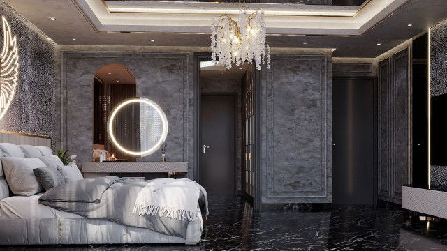 The picture depicts an ultra-modern living room, with a white L-shaped couch and a black ottoman. The walls are a light grey, with modern shelving on either side of the room. There is a glass top table in the middle of the room, and the floor is covered in a dark coloured carpet. A large painting hangs on the far wall, and there are two pendant lights hanging from the ceiling.