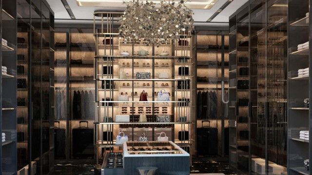 This image shows a living room featuring a modern and luxurious design. The room has a stylish gray color palette, featuring a large sectional sofa and accent chairs in shades of gray, a round black coffee table, and a rectangular rug with a subtle geometric pattern. On the walls, there are several pieces of abstract art in shades of blue and gray. The room is illuminated by multiple recessed lights, and a large crystal chandelier hangs from the ceiling, adding a touch of glamour to the space.