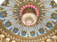 The picture shows a woman in a bright green dress walking down a cobblestone street in Portugal. She appears to be carrying bags and looking straight ahead. The buildings along the street are brightly painted in various colors, with red tiling on the roofs, and there are beautiful hanging flowers in view.